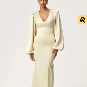 Champagnefärgad långklänning med v-ringning - Elegant långklänning i champagnefärgad satin med djup v-ringning och långa puffärmar. Klänningen har figurnära passform upptill och vida ärmar som ger en lyxig känsla. Perfekt för speciella tillfällen.