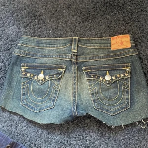 True Religion jeansshorts med nitar - Snygga jeansshorts från True Religion i mörkblå denim med slitna kanter och coola nitar på fickorna. Klassiska kontrastsömmar och detaljerad brodyr på bakfickorna. Perfekt för dig som vill sticka ut med en edgy streetstil.