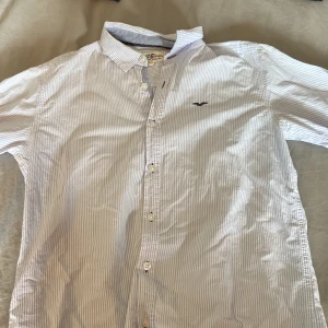 Randig skjorta från Hampton Republic - Snygg skjorta från Hampton Republic i ljusblått och vitt med tunna ränder. Klassisk krage, knäppning framtill och broderad logga på bröstet. Skjortan är långärmad och tillverkad i mjuk bomull, perfekt för en clean och fräsch look. Storlek: 170, skick: 8/10, kontakta vid frågor!