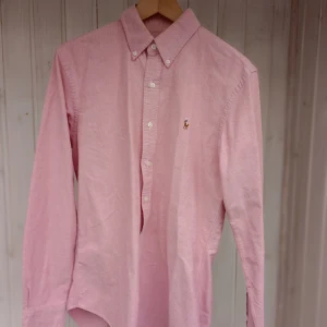 Rosa skjorta Polo Ralph Lauren M - Snygg rosa skjorta från Polo Ralph Lauren i slim fit-modell. Skjortan har klassisk krage, långa ärmar och knappar framtill. Broderad logga på bröstet. Perfekt för dig som gillar stilrena och tidlösa plagg.