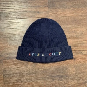 Lyle&scott Mössa - Mörkblå Lyle & Scott-mössa i ribbstickat tyg med färgad logotyp framtill. Stilren, bekväm och perfekt för kallare dagar.