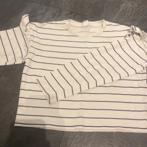 Randig långärmad tröja från Lindex - Vit långärmad tröja från Lindex med tunna marinblå ränder. Tröjan har en klassisk rund halsringning och är gjord i mjuk bomull. Perfekt för en chill och stilren look, funkar året runt och är lätt att matcha med jeans eller kjol.