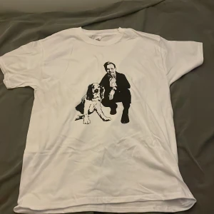Vit t-shirt med tryck från Fruit of the Loom - Vit t-shirt från Fruit of the Loom i bomull med svartvit tryck av en person i kostym och en hund på framsidan. Klassisk passform med rund hals och korta ärmar. Perfekt för dig som gillar unika motiv och enkel stil.