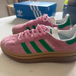 Adidas Gazelle rosa/gröna sneakers - Adidas Gazelle sneakers i rosa mocka med gröna ränder och vit häl. Skorna har en chunky brun gummisula och klassisk snörning. Ikonisk retrostil med rund tå och gröna detaljer på insidan. Perfekta för dig som vill sticka ut med färg och stil.             Skorna är oanvända. 