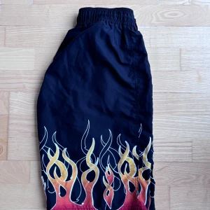 Svarta flame shorts med snörning - Svarta shorts med elastisk midja och snörning, dekorerade med eldflammor i gult, orange och rött längs benen. Insidan har gul mesh för extra komfort. Perfekta för dig som vill sticka ut med en cool och edgy stil.