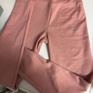 Rosa ribbade leggings från Nike - Säljer ett par snygga rosa leggings från Nike med diskret ribbad struktur och klassisk Swoosh-logga i silver på benet. Perfekta för träning eller chill, med mjukt och stretchigt material som sitter skönt hela dagen. Passar även xxs. Se tröja till i separat annons.