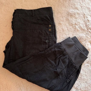 Svarta byxor från Vero Moda - Svarta byxor från Vero Moda med knäppning framtill och muddar vid bensluten. Byxorna har sidfickor med knappdetaljer och en avslappnad passform. Perfekta för en chill och stilren look.