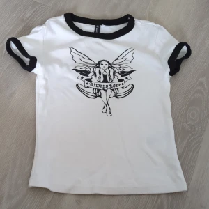 Vit och svart t-shirt med fe-tryck H&M - Vit t-shirt från H&M Divided med svarta kontrastkanter på ärmar och hals. Tryck framtill med en fe och texten 'Always Love' i svart. Modellen är kortärmad och i 100% bomull, perfekt för en cool och avslappnad stil.