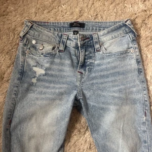 True Religion ljusa bootcut jeans - Säljer ett par ljusblå bootcut jeans från True Religion med slitna detaljer och snygga röda sömmar. Klassiska femficksmodellen med logga på bakfickan och coola knappar. Perfekta för dig som gillar en avslappnad och trendig look. Använda  3 gånger så i fint skick!