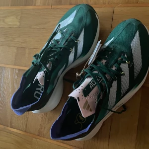 Adidas Adizero Adios Pro 3 löparskor - Gröna Adidas Adizero Adios Pro 3 med vita och svarta detaljer. Skorna har ventilerande mesh-ovandel, snörning och en tjock, responsiv sula för maxad löpkänsla. Lila insida och perforerad plös i ljusrosa. Perfekta för dig som vill springa snabbt och snyggt. De är samples och aldrig använda pga fel storlek. 