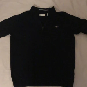 Svart stickad half zip tröja GANT - Svart stickad tröja från GANT med broderad logga på bröstet. Tröjan har half zip-dragkedja och hög krage, perfekt för lager-på-lager. Klassisk och stilren modell som funkar till det mesta. Mjuk och skön känsla.