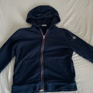 Mörkblå hoodie från Moncler - Säljer en mörkblå hoodie från Moncler i 100% bomull. Tröjan har hel dragkedja med röd-vita detaljer, huva och två fickor framtill. Moncler-logga sitter på vänster ärm. Perfekt för dig som gillar stilrena och exklusiva plagg.
