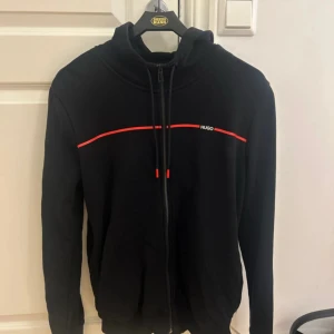 Svart hoodie från Hugo Boss med röd detalj - Svart hoodie från Hugo Boss med röd linje över bröstet och diskret HUGO-logga. Tröjan är i ny skick och använd någon gång! Hör av er vid frågor😁