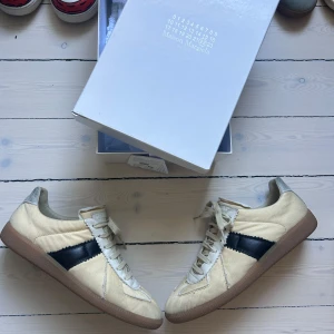 Maison Margiela sneakers med svart detalj - Unika sneakers från Maison Margiela i beige canvas med vita och svarta detaljer samt bruna gummisulor. Skorna har snörning, rund tå och en cool kontrast med svart sidopanel. Perfekta för dig som gillar stilrena men ändå edgy sneakers.