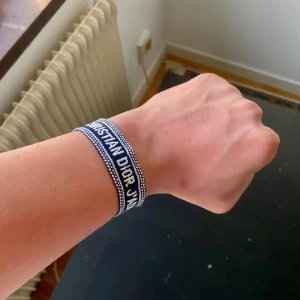 Christian Dior J'Adior armband blå - Snyggt armband från Christian Dior med texten 'J'Adior' broderad i vitt på blå bakgrund. Armbandet är gjort i tyg och har knytning med tofsar i ändarna. Perfekt accessoar för att lyfta din outfit med en lyxig touch.