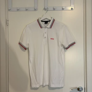 Vit BOSS pikétröja med röd logga - Vit pikétröja från BOSS med rosa logga på bröstet. Krage och ärmslut har snygga detaljer i grått och rosa . Klassisk passform och tillverkad i bomull, perfekt för en clean och stilren look.