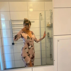 Ljusrosa blommig långklänning - Supersnygg långklänning i ljusrosa med blommigt mönster och v-ringning. Klänningen har långa ärmar och en slits framtill som ger en extra touch. Perfekt för dig som vill sticka ut med en feminin och trendig look.