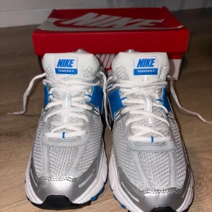 Nike Vomero 5 silver/blå sneakers - Nike Vomero 5 sneakers i silver, vitt och blått med mesh och syntetpaneler. Skorna har snörning, svart swoosh och detaljer i blått på sidorna och hälen. Perfekta för dig som gillar sportig och modern stil.