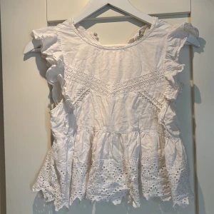 Vit blus med volang och spetsdetaljer - Superfin vit blus med volangärmar och broderade spetsdetaljer. Toppen har rund hals och broderade hålmönster längs fållen. 