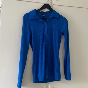 Blå ribbad skjorta från Gina Tricot - Snygg blå skjorta från Gina Tricot med ribbad struktur och klassisk krage. Skjortan har lång ärm, knappar framtill och en figurnära passform. Perfekt att styla med jeans eller kjol för en trendig look.