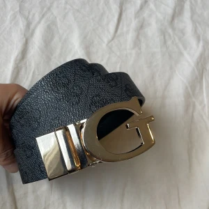 Svart Guess bälte med G-spänne - Snyggt svart bälte från Guess med diskret mönster och ett stort, glänsande G-format spänne i guld. Bältet är gjort i skinn och har en stilren look som lyfter vilken outfit som helst. Perfekt för dig som gillar statement-accessoarer.