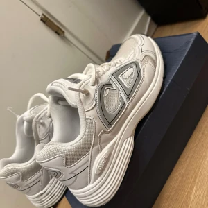Vita Dior b30 skor - Snygga vita sneakers från Dior med stora CD-loggor på sidorna och hälen. Skorna har en chunky sula, meshpaneler för extra andningsförmåga och klassisk snörning. Perfekta för dig som vill sticka ut med exklusiva och trendiga sneakers.