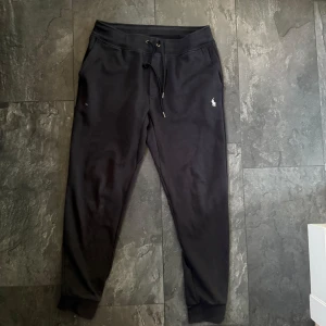 Svarta mjukisbyxor från Ralph Lauren - Svarta mjukisbyxor från Ralph Lauren med klassisk vit logga på vänster ben. Byxorna har snörning i midjan, muddar vid bensluten och är tillverkade i mjukt bomullsmaterial. Perfekta för chill dagar eller när du vill vara bekväm men ändå se snygg ut.