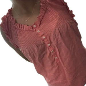 Rosa blus med volang  - Supersöt rosa blus med volangkant vid halsringningen och ärmarna. Smockad upptill och knäppning framtill med små vita knappar. Storlek S
