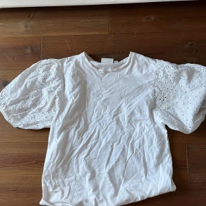 Vit blus med puffärmar och broderi - Supersöt vit blus med korta puffärmar i broderad spets och rund halsringning. Ärmarna har fina hålbroderier som ger en romantisk känsla. Toppen är i mjuk bomull och har en avslappnad passform, perfekt till jeans eller kjol.