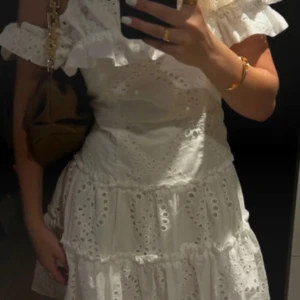 Vit broderad kortklänning med volang - Superfin vit kortklänning med broderade detaljer och volang vid axlarna. Klänningen har offshoulder-modell och är tillverkad i bomull, perfekt för sommarens alla tillfällen. Den har en luftig passform och romantisk känsla.