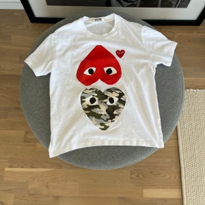 Vit Comme des Garçons Play t-shirt - Vit t-shirt från Comme des Garçons Play med två stora hjärttryck på framsidan, ett rött och ett i camouflage. Klassisk passform och korta ärmar. Ikonisk design som sticker ut och passar perfekt till jeans eller shorts. Storlek S men sitter aningen kort hade passat någon som är 172 och neråt. Tröjan är använd en gång
