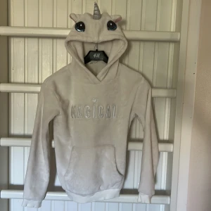 Magisk enhörningshoodie från H&M - Supermjuk hoodie från H&M i offwhite fleece med broderad text 'MAGICAL' på bröstet. Huvan har söta enhörningsdetaljer med horn, öron och stora glittriga ögon. Perfekt för dig som älskar gulliga och unika plagg!