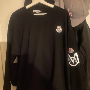Svart långärmad Moncler tröja - Svart långärmad tröja från Moncler med klassisk rund halsringning. Tröjan har Moncler-logga på bröstet och ärmen samt en stor vit M-detalj på ena ärmen. Snygg och stilren design som passar perfekt till streetwear. Använd 2 gånger bara. 10/10 i skick