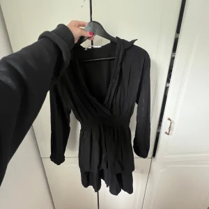 Svart omlottklänning från Zara XS - Supersnygg svart kortklänning från Zara i omlottmodell med v-ringning och markerad midja. Klänningen har långa ärmar och lätt rynkad kjol, vilket ger en chill men ändå stilren vibe. Perfekt för dig som gillar enkel men trendig stil.