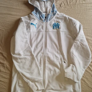 Vit hoodie från Puma x Marseille - Vit hoodie med dragkedja från Puma och Olympique de Marseille. Har blå detaljer, klubbmärke på bröstet och randigt foder i huvan. Väldigt unik då det är en gammal modell, perfekt för franska stilen.