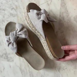 Beige sandaler med rosett och platåsula - Snygga beige sandaler med bred rosett framtill och flätad platåsula i espadrillestil. Perfekta för sommaren och ger en chill vibe till din outfit. Mjukt tyg upptill och bekväm fotbädd.