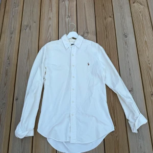 Vit skjorta från Polo Ralph Lauren M - Klassisk vit skjorta från Polo Ralph Lauren i slim fit med broderad logga på bröstet. Skjortan har lång ärm, knappar framtill och är tillverkad i mjuk bomull. Perfekt för en clean och stilren look.