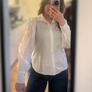 Vit skjorta med puffärm  - Klassisk vit skjorta med snygga puffärmar och markerade manschetter. Skjortan har krage och knäppning framtill, vilket ger en clean och stilren look. Perfekt att styla med jeans eller kjol för en trendig vibe.