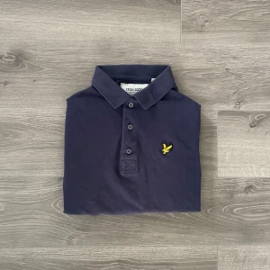 Mörkblå pikétröja från Lyle & Scott - Snygg mörkblå pikétröja från Lyle & Scott i ekologisk bomull. Klassisk krage, tre knappar och ikonisk gul logga på bröstet. Perfekt för en clean och stilren look. Skön passform och mjukt material. Hör av dig vid funderingar😊