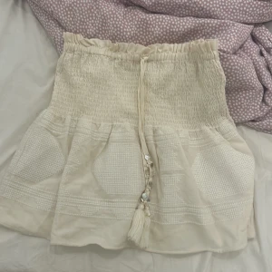 Kjol från Zara - Superfin beige kortkjol från Zara med smockad midja och dekorativ snörning med pärlor och tofsar. Kjolen har broderade detaljer och är gjord i ett mjukt bomullstyg. Perfekt för sommarens alla tillfällen!