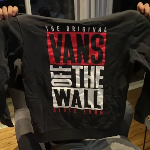 Svart hoodie från Vans med tryck - Svart hoodie från Vans med stort rött och vitt 'Vans Off The Wall' tryck på baksidan och mindre Vans-logga på framsidan. Klassisk känguruficka framtill och huva. Perfekt för dig som gillar streetwear och vill sticka ut med en fet logga.