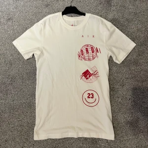 Vit Jordan t-shirt med röd print - Snygg vit t-shirt från Jordan med röda tryck och coola grafiska detaljer på framsidan, inklusive texten 'AIR JORDAN' och siffran 23. Klassisk passform och rund hals. Perfekt för dig som gillar streetwear och sportig stil.