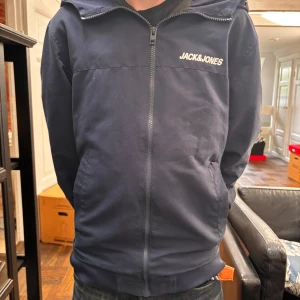 Mörkblå vindjacka från Jack & Jones - Säljer en snygg mörkblå vindjacka från Jack & Jones med huva och dragkedja hela vägen. priset är ej hugget vid sten vid snabb affär. Jag på bilden är 170 om ni vill ha referens 