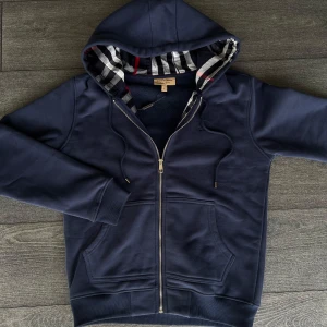 Marinblå burberry zip - Snygg marinblå hoodie från Burberry med dragkedja och klassiskt rutigt foder i huvan. Tröjan har snörning vid huvan, två fickor fram och broderad logga på bröstet. Perfekt för dig som vill ha en stilren och lyxig look.