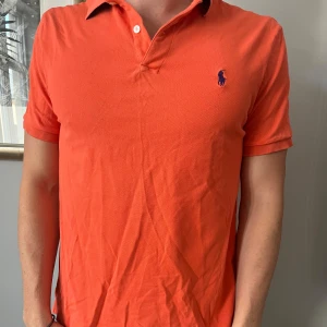 Orange pikétröja från Ralph Lauren - Säljer en orange pikétröja från Ralph Lauren med klassisk krage och två knappar. Tröjan har korta ärmar och den ikoniska broderade loggan i mörkblått på bröstet. Tillverkad i bomull för en skön och luftig känsla. skick 7/10, Stolek M 