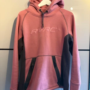 Rosa hoodie från RevolutionRace - Säljer en rosa hoodie från RevolutionRace med mörkgrå detaljer längs sidorna och ärmarna och ficka med dragkedja framtill. 