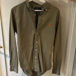 Olivgrön skjorta från Ralph Lauren - Säljer en olivgrön långärmad skjorta från Ralph Lauren i slim fit. Skjortan har knappar framtill och en broderad logotyp i rött på bröstet. Perfekt för en stilren look. Nypris 1700 kr 