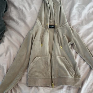 Beige velour hoodie från Juicy Couture - Säljer en beige hoodie i mjuk velour från Juicy Couture. Tröjan har dragkedja, huva med snören och två fickor framtill. Broderad JC-logga på bröstet och guldfärgade detaljer på snörändarna.