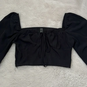 Svart croppad topp med puffärmar från SHEIN - Trendig svart croppad topp från SHEIN med långa puffärmar och knytning framtill. Toppen har en fyrkantig urringning och är tillverkad i ett lätt, syntetiskt material..