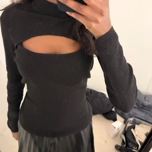 Svart ribbad långärmad topp med cut-out - Säljer en svart ribbad långärmad topp med en snygg cut-out detalj över bröstet. Toppen har en tight passform och hög hals, perfekt för att skapa en trendig look.